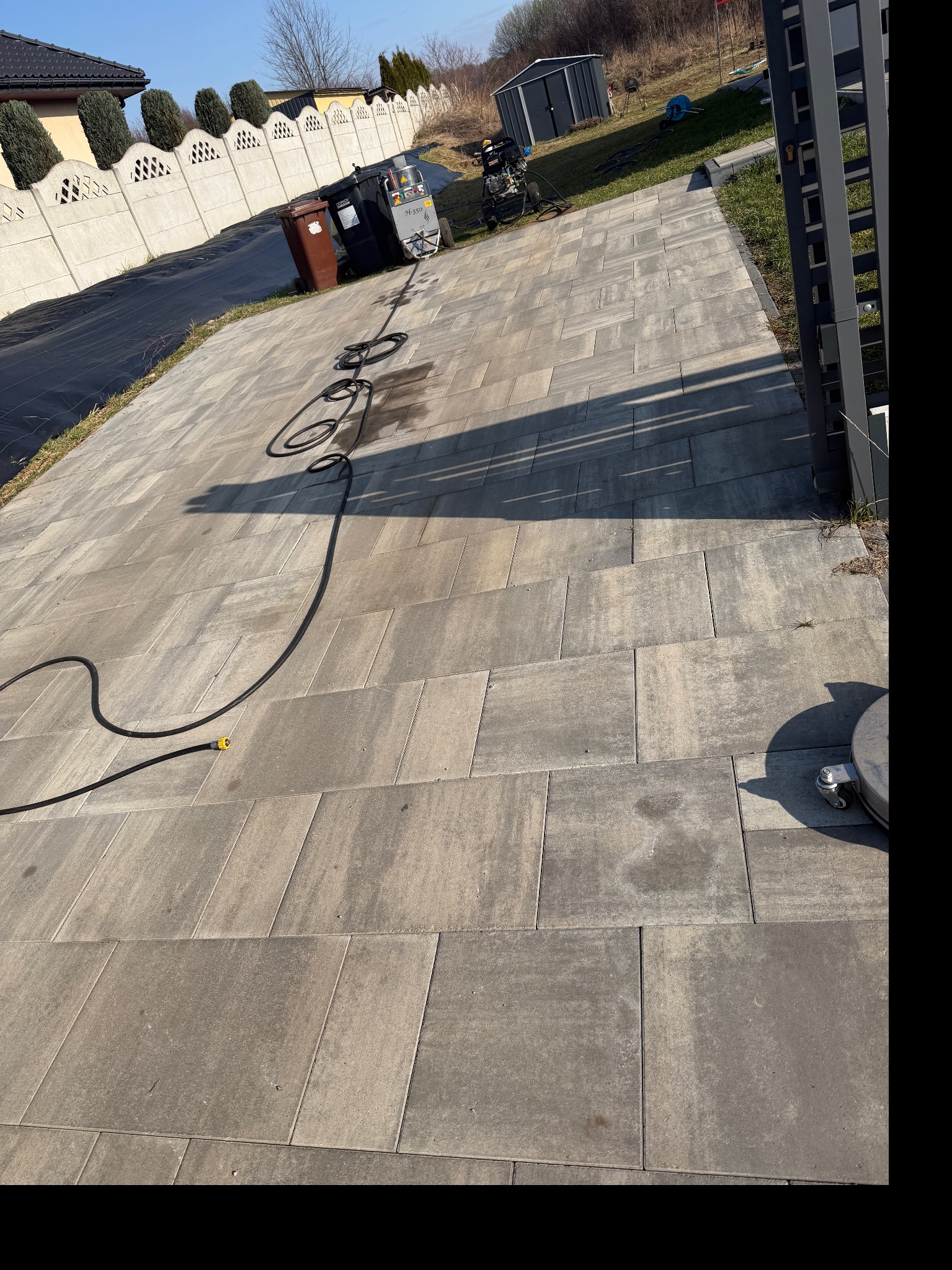 Kostka brukowa po czyszczeniu w Chrzanowie. Widoczny wąż ciśnieniowy i urządzenie czyszczące. Czysta, szara kostka na podjeździe, w tle ogrodzenie i zieleń.