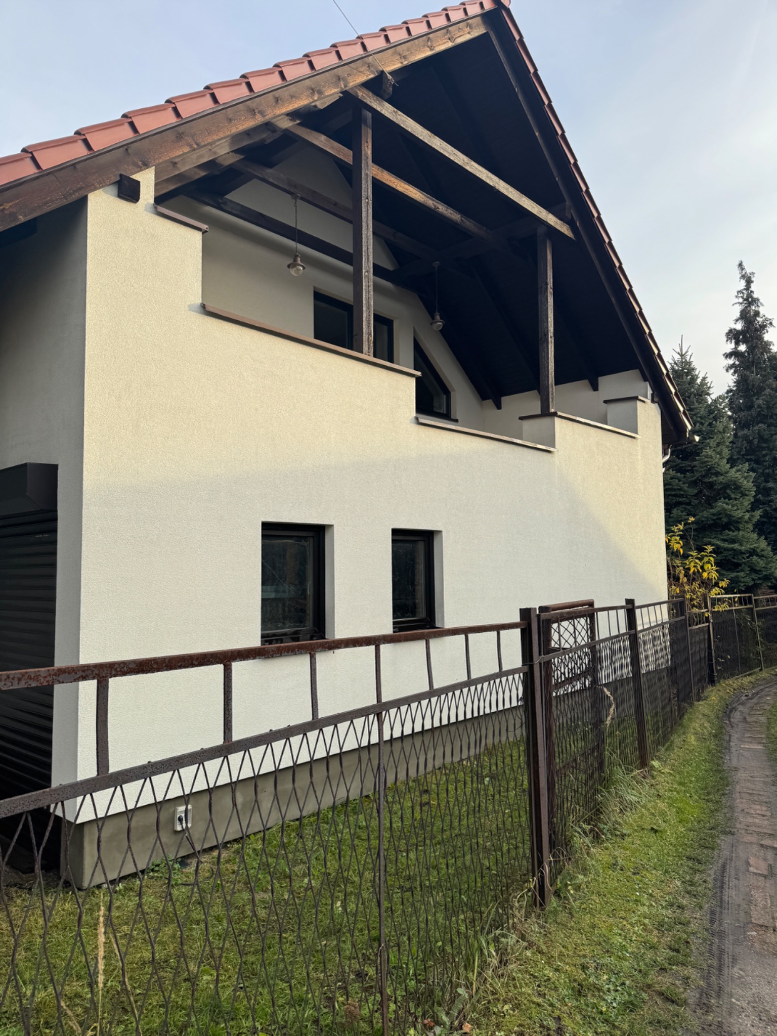Dwupiętrowy dom z białymi ścianami i drewnianym balkonem, widziany zza metalowego ogrodzenia. Widoczne okna i fragment dachu. Tło stanowią drzewa.