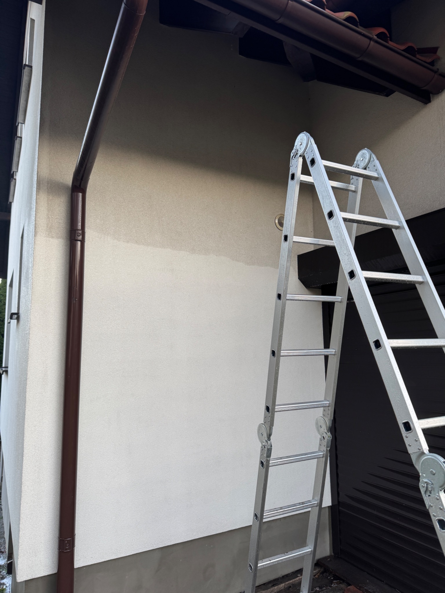 Fragment elewacji domu po częściowym umyciu, z widoczną różnicą w kolorze. Aluminiowa drabina oparta o ścianę, brązowa rynna wzdłuż krawędzi. Ujęcie z dołu.