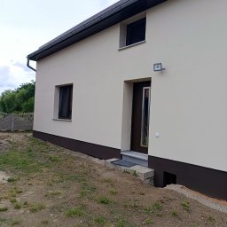 B&K FLEX SP. Z O.O. - Elewacja domu po remoncie: kremowa ściana, brązowy cokół, nowe okna i drzwi. Widoczny fragment ogrodu i ogrodzenia. Dom z oświetleniem zewnętrznym.