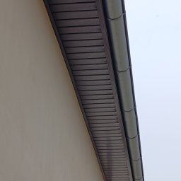 B&K FLEX SP. Z O.O. - Brązowa podbitka dachowa z rynną, zamontowana przy beżowej ścianie budynku. Ujęcie z dołu, widoczne łączenia elementów podbitki.