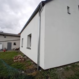 B&K FLEX SP. Z O.O. - Świeżo pomalowana, biała elewacja budynku z rynną, widoczna z zewnątrz w pochmurny dzień. W tle drugi budynek i elementy placu budowy.