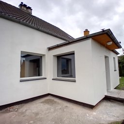 B&K FLEX SP. Z O.O. - Biała elewacja domu jednorodzinnego z dwoma oknami i dobudówką, widok z zewnątrz w pochmurny dzień. Widoczne detale wykończenia i fragment dachu.