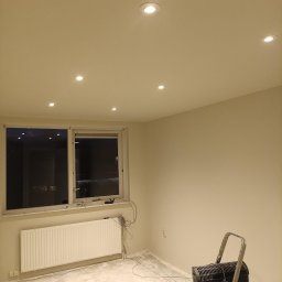 B&K FLEX SP. Z O.O. - Wnętrze po malowaniu ścian na jasny kolor, widoczne nowe oświetlenie sufitowe, grzejnik, okno, drabina i odkurzacz przemysłowy. Przygotowanie do dalszych prac wykończeniowych.