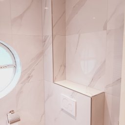 B&K FLEX SP. Z O.O. - Wykończona łazienka z okrągłym oknem i białymi płytkami imitującymi marmur. Widoczny podtynkowy system WC i półka z tego samego materiału.