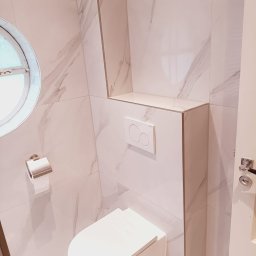 B&K FLEX SP. Z O.O. - Nowoczesna toaleta z marmurowymi płytkami, okrągłym oknem i podtynkową spłuczką. Minimalistyczny design, jasne barwy, widoczny uchwyt na papier toaletowy i fragment drzwi.