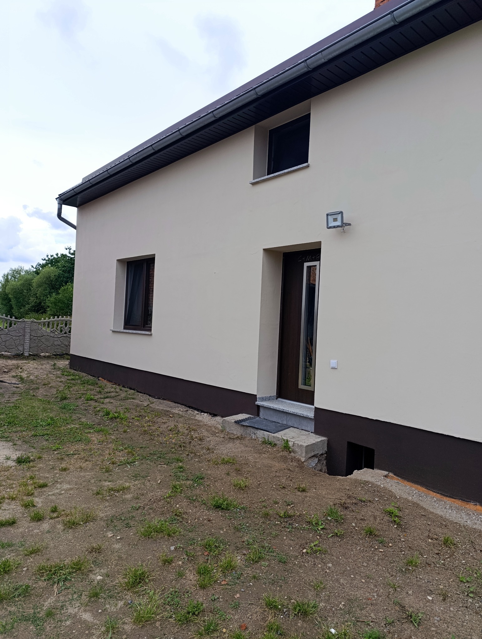 Elewacja domu po remoncie: kremowa ściana, brązowy cokół, nowe okna i drzwi. Widoczny fragment ogrodu i ogrodzenia. Dom z oświetleniem zewnętrznym.