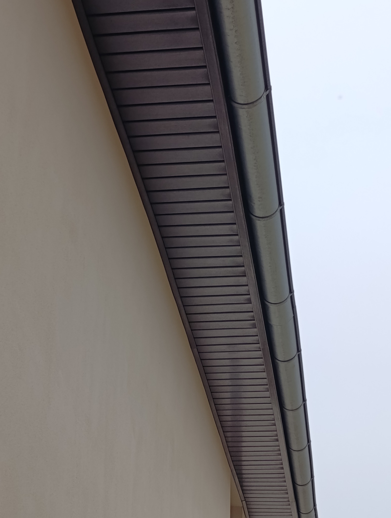 Brązowa podbitka dachowa z rynną, zamontowana przy beżowej ścianie budynku. Ujęcie z dołu, widoczne łączenia elementów podbitki.