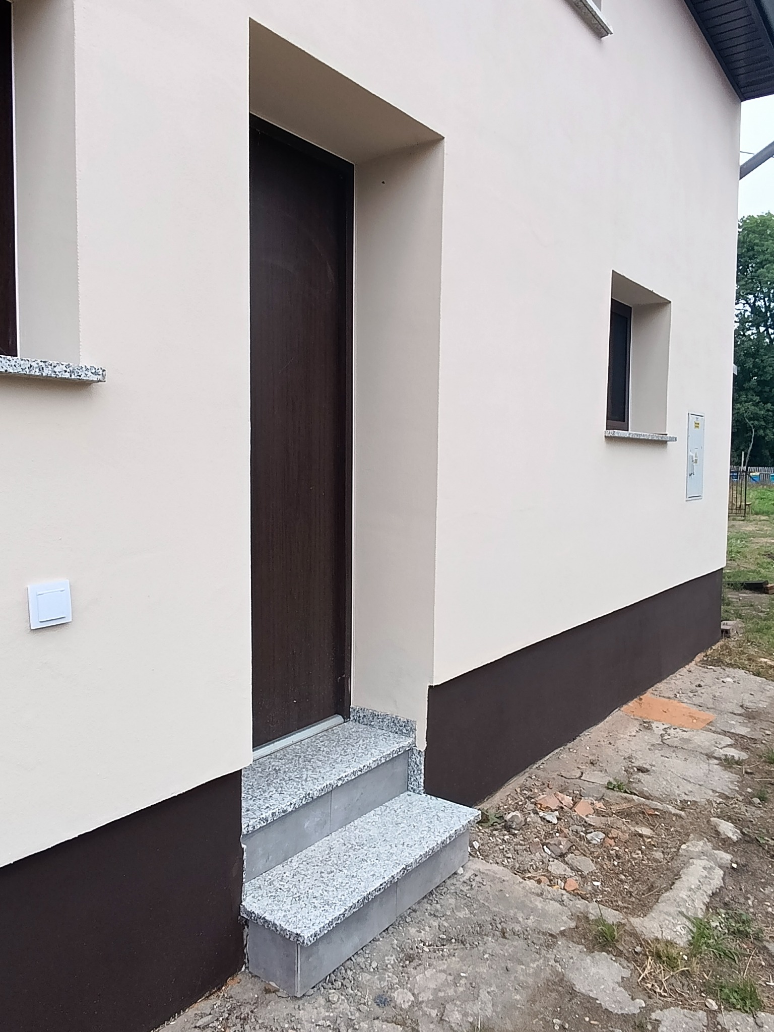 Elewacja domu po malowaniu w odcieniach beżu i brązu, z granitowymi schodami wejściowymi. Widoczny fragment okna i drzwi wejściowych. Detal wykończenia.