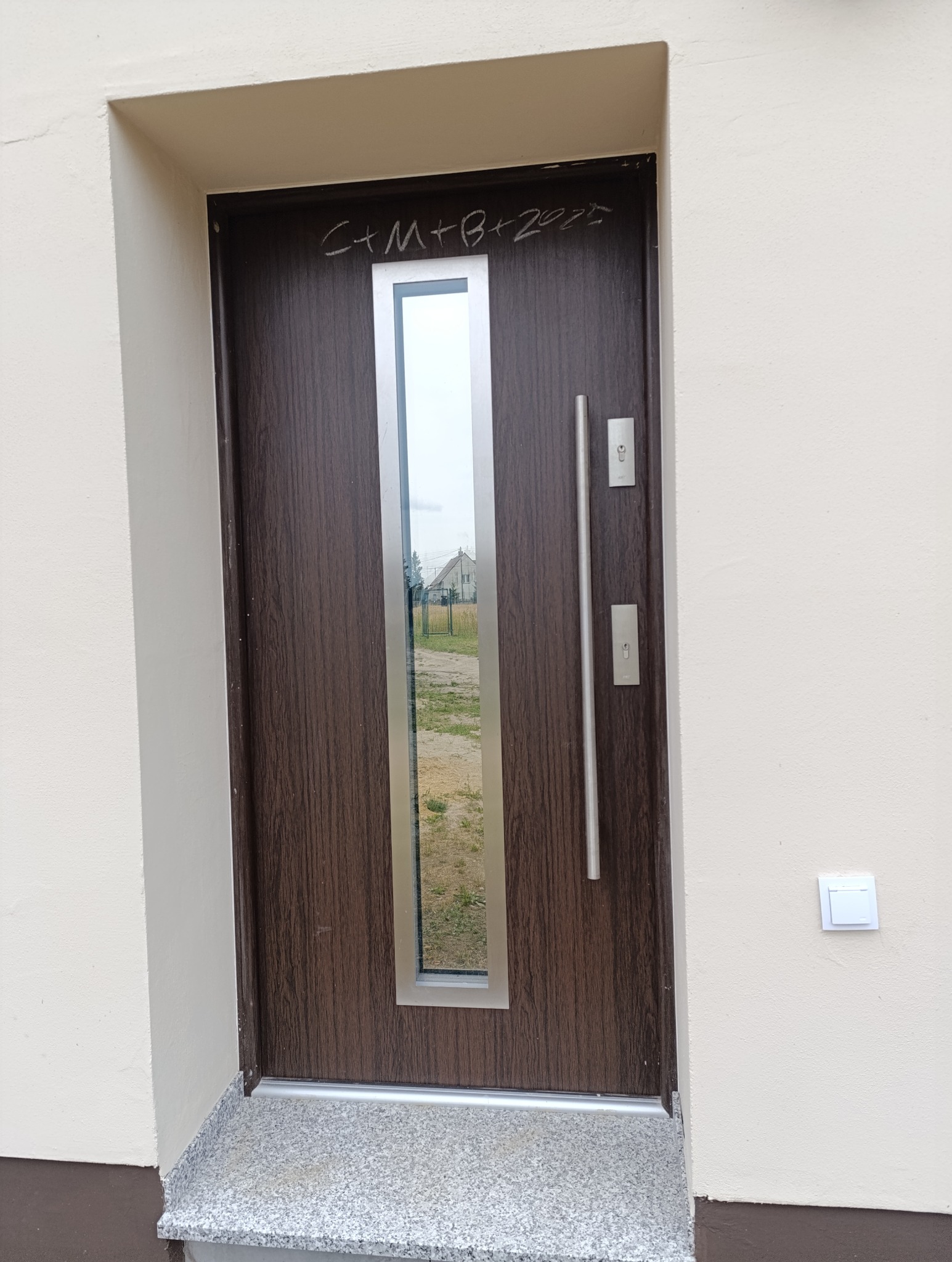 Zamknięte, brązowe drzwi wejściowe z długim, srebrnym uchwytem i szybą, osadzone w beżowej ścianie. Nad drzwiami napis 'C+M+B+2023'.
