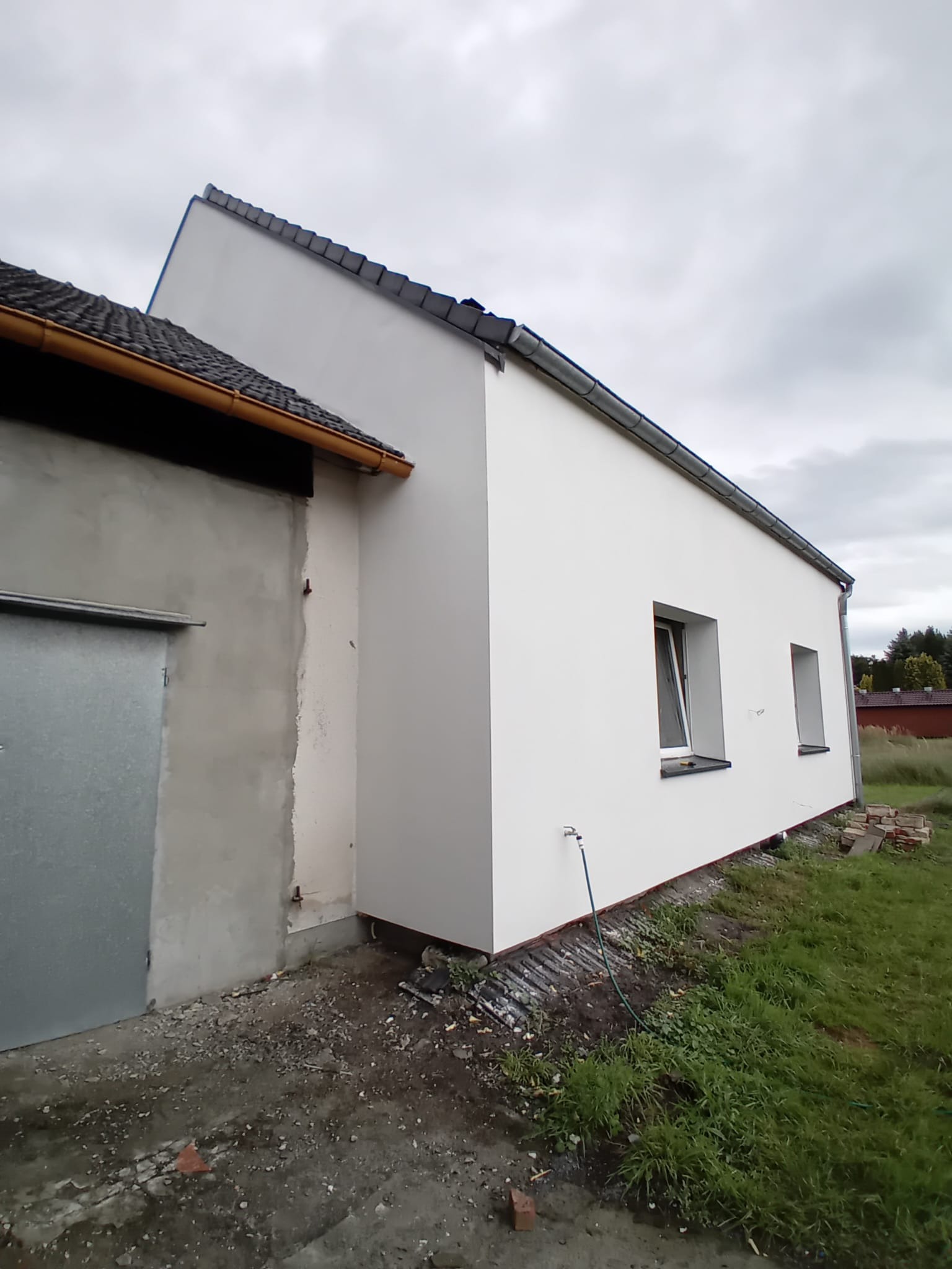 Boczny widok otynkowanego budynku z dwoma oknami, z fragmentem elewacji w stanie surowym. Widoczne elementy dachu i otoczenie budynku.