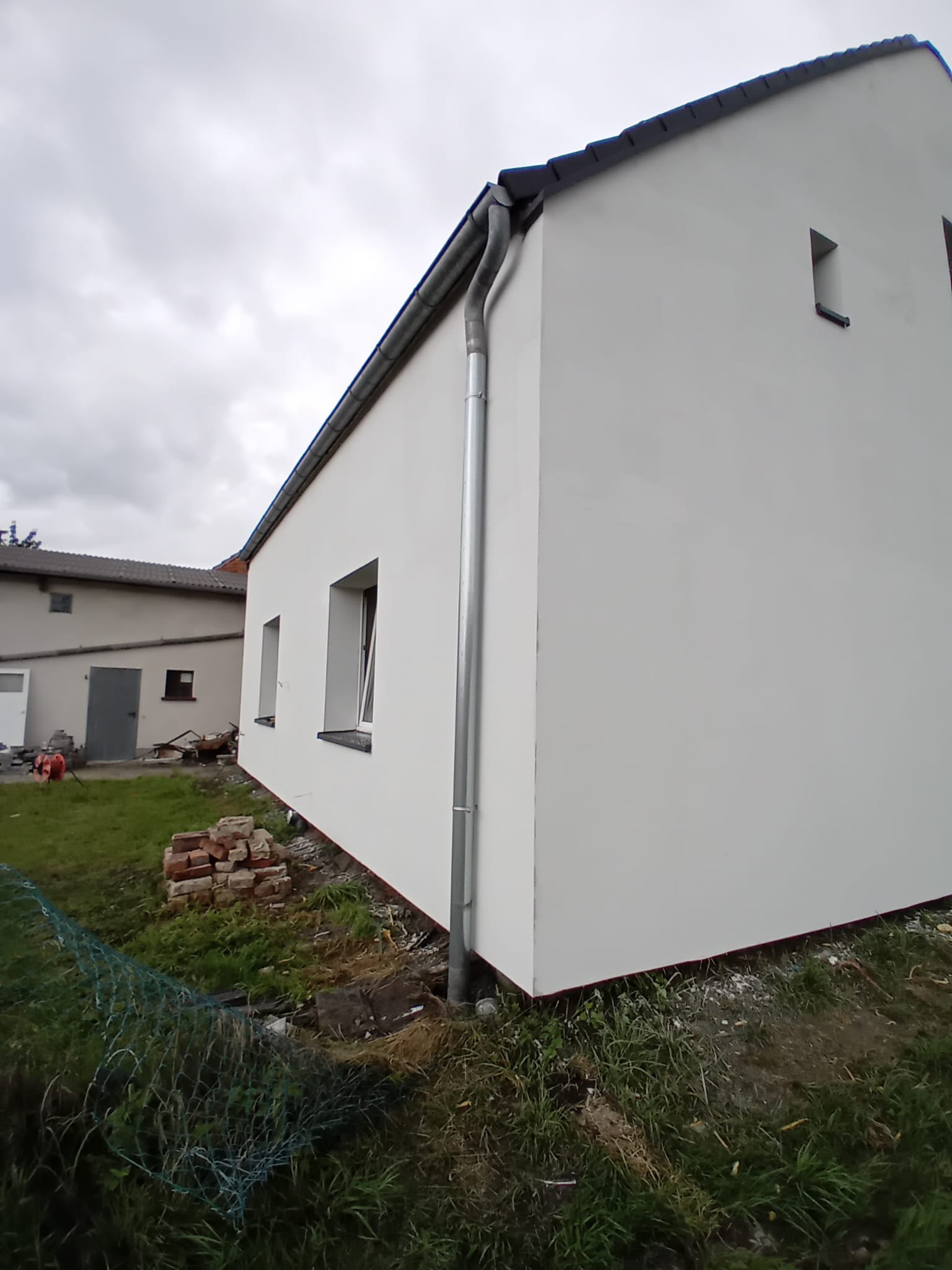 Świeżo pomalowana, biała elewacja budynku z rynną, widoczna z zewnątrz w pochmurny dzień. W tle drugi budynek i elementy placu budowy.