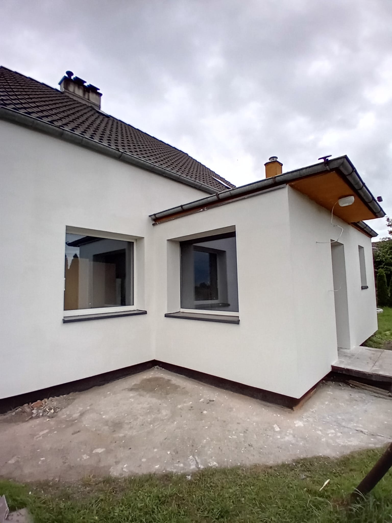 Biała elewacja domu jednorodzinnego z dwoma oknami i dobudówką, widok z zewnątrz w pochmurny dzień. Widoczne detale wykończenia i fragment dachu.