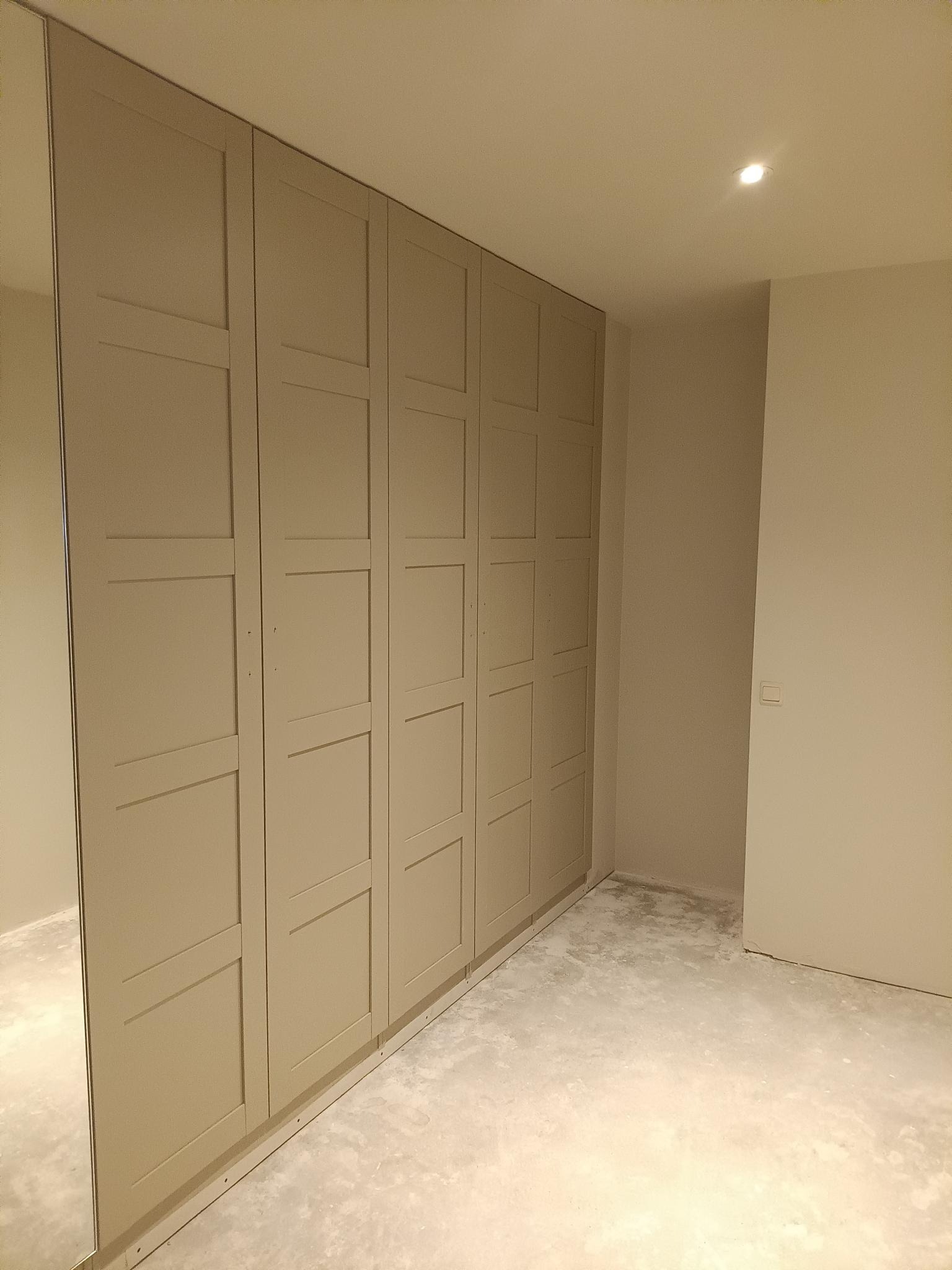 Beżowa zabudowa szafy wnękowej z lustrem na boku, widoczna w surowym stanie pomieszczenia. Minimalistyczny design, ciepłe oświetlenie sufitowe.