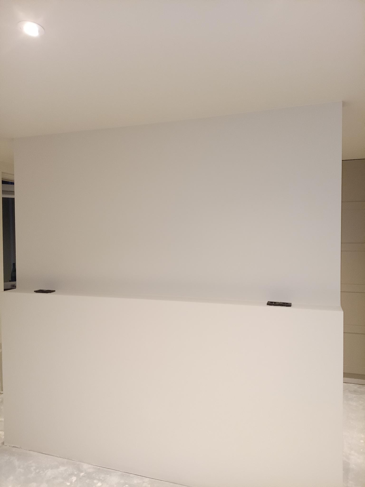 Gładka, świeżo pomalowana ściana w jasnym odcieniu bieli, z widocznymi śladami po taśmie malarskiej na dolnej części. Minimalistyczne wnętrze w trakcie wykończenia.