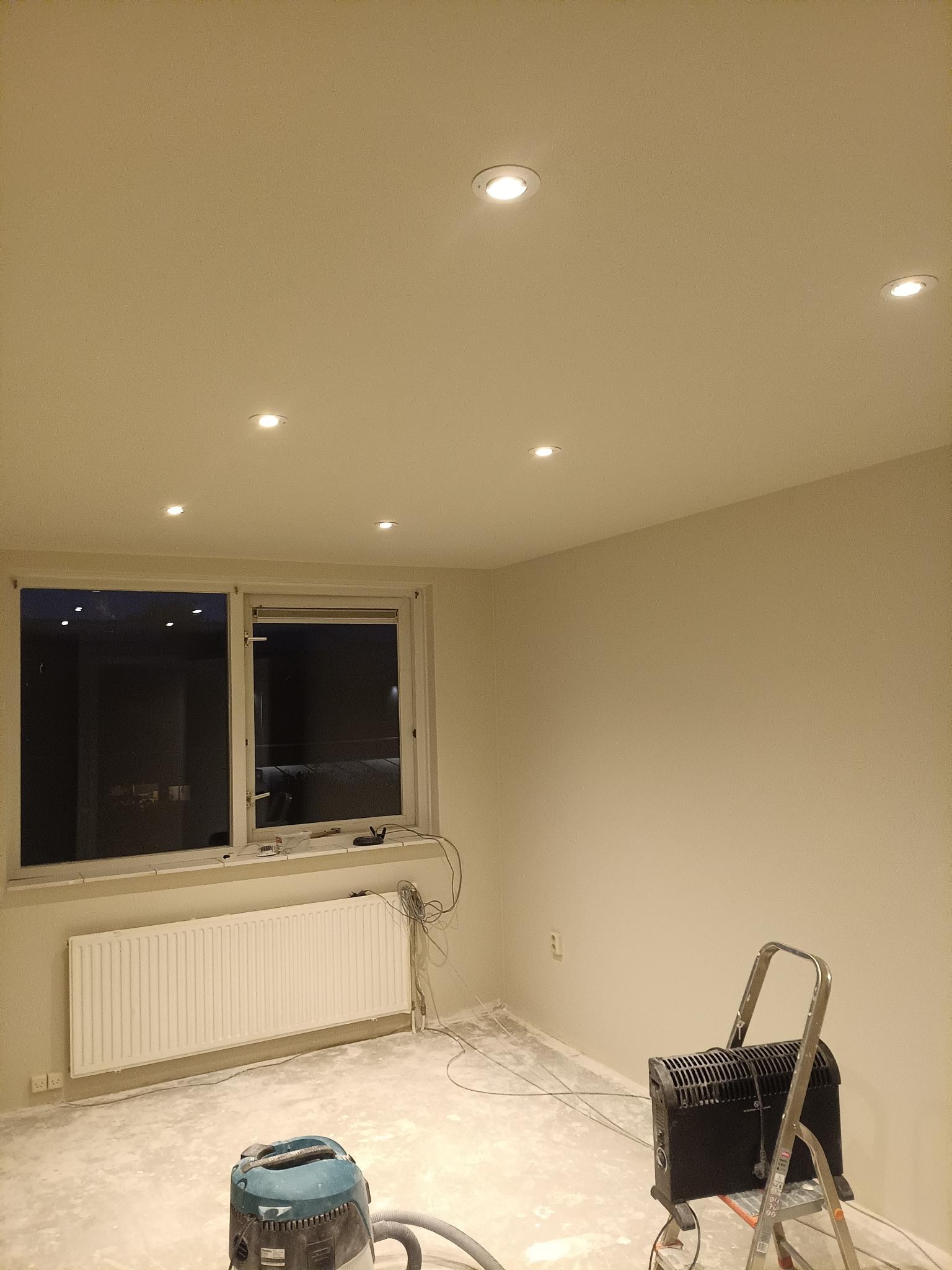 Wnętrze po malowaniu ścian na jasny kolor, widoczne nowe oświetlenie sufitowe, grzejnik, okno, drabina i odkurzacz przemysłowy. Przygotowanie do dalszych prac wykończeniowych.