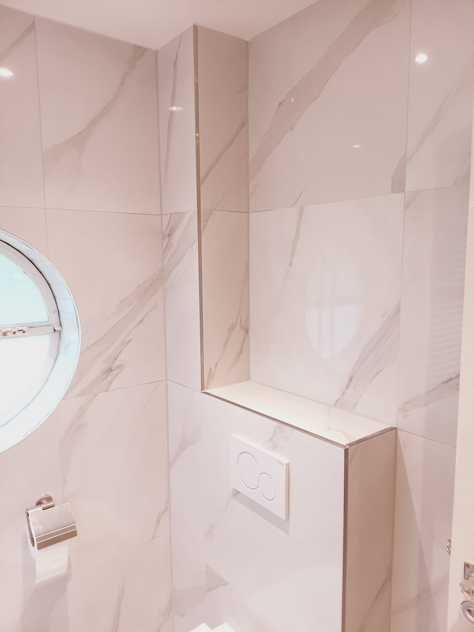 Wykończona łazienka z okrągłym oknem i białymi płytkami imitującymi marmur. Widoczny podtynkowy system WC i półka z tego samego materiału.