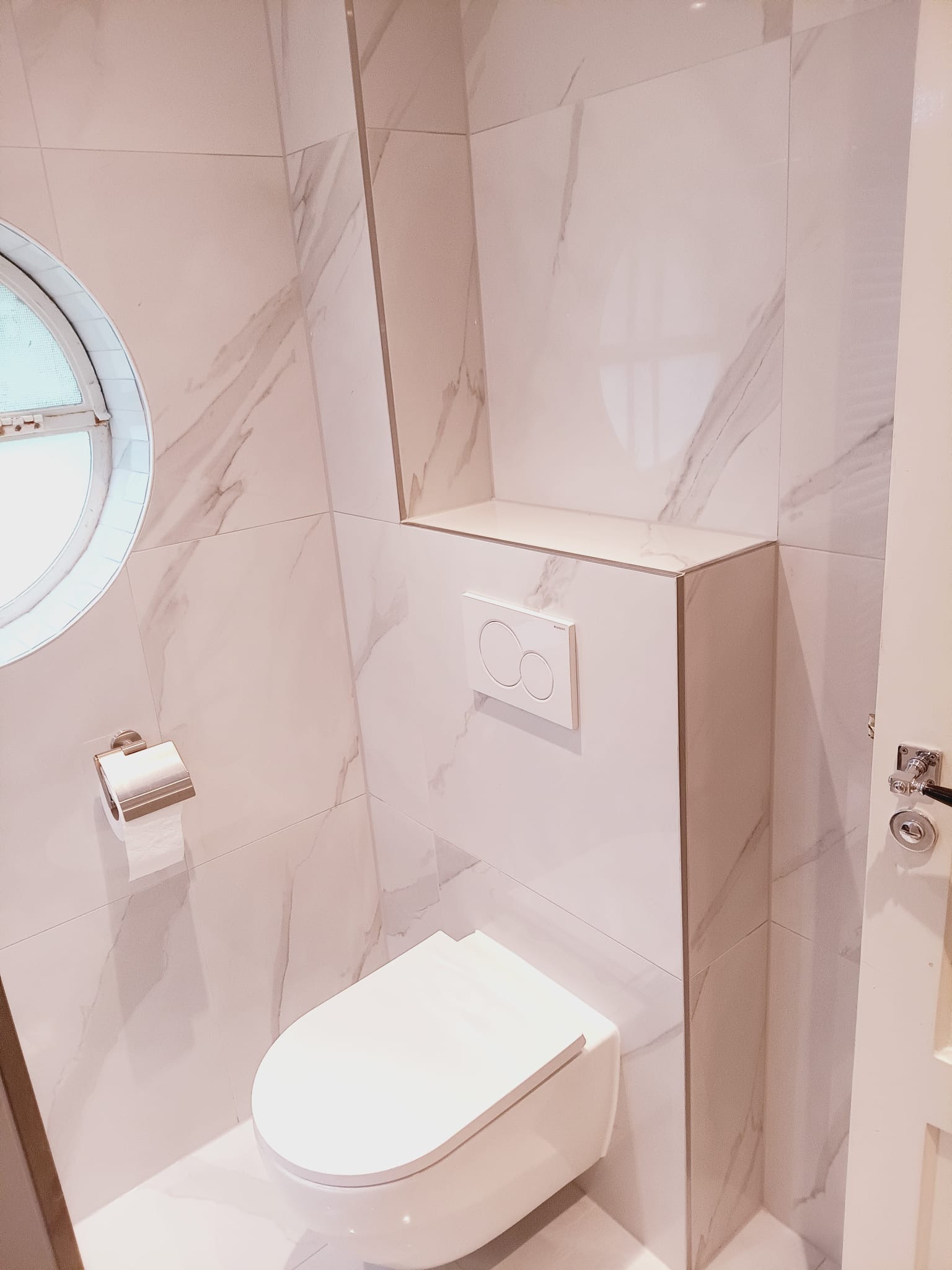 Nowoczesna toaleta z marmurowymi płytkami, okrągłym oknem i podtynkową spłuczką. Minimalistyczny design, jasne barwy, widoczny uchwyt na papier toaletowy i fragment drzwi.