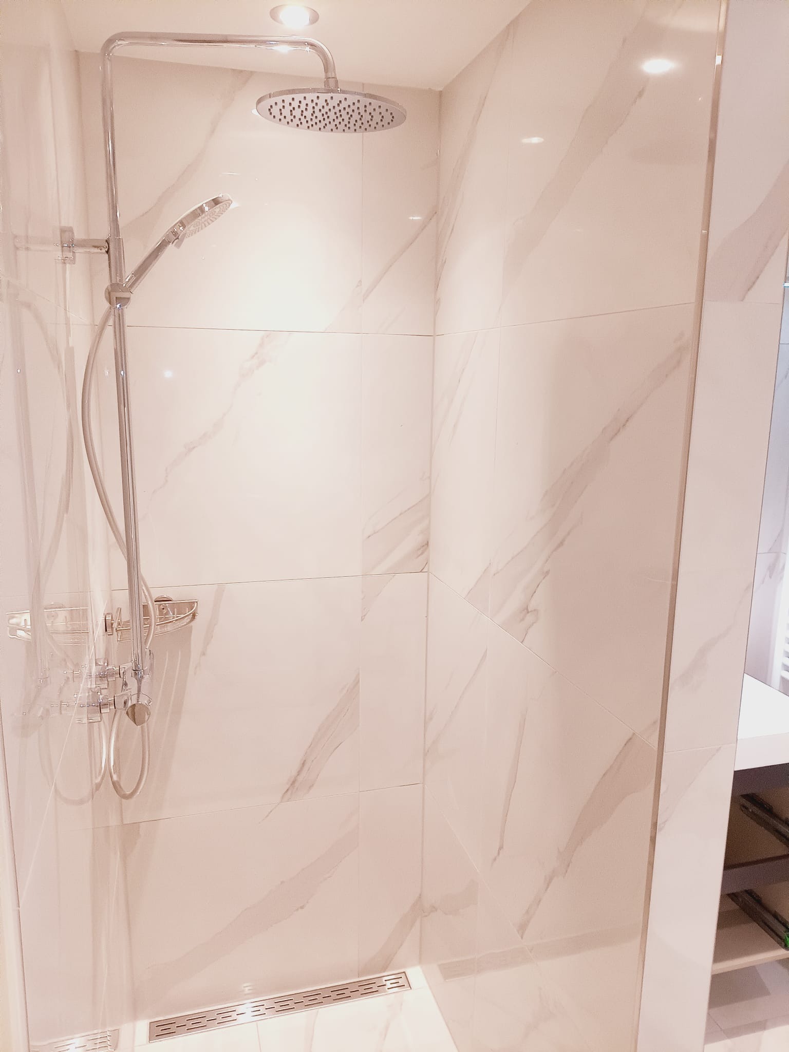 Nowoczesna kabina prysznicowa z deszczownicą i marmurowymi płytkami w odcieniach bieli i beżu. Widoczny odpływ liniowy i półka na kosmetyki.