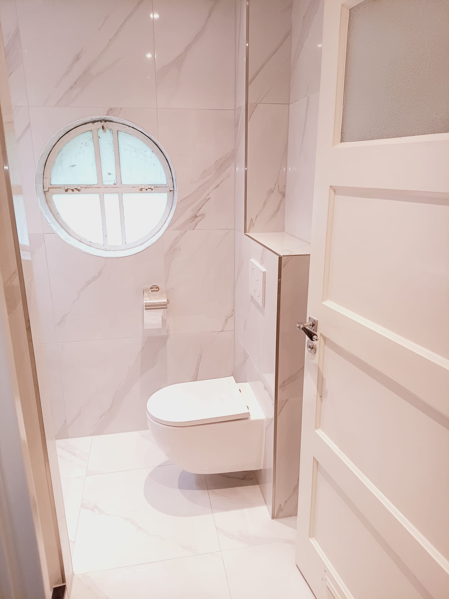 Wykończona łazienka w bieli z marmurowym wzorem, okrągłe okno z kratką, podwieszana toaleta i częściowo otwarte białe drzwi z przeszkleniem.