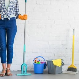 PROFESJONALNE SPRZĄTANIE MAK-MAR Aldona Kawalec - Osoba w jeansach i rękawicach z mopem, wiadrem i zmiotką na tle białej ściany z cegieł. Widoczne środki czystości. Ujęcie od pasa w dół.