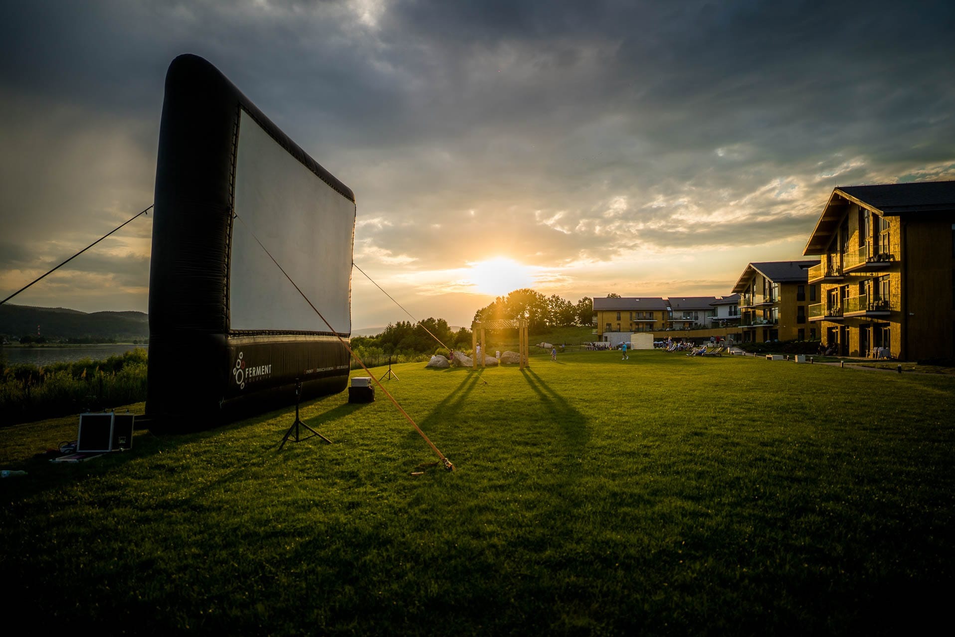 Plenerowe kino z dmuchanym ekranem na zielonej trawie o zmierzchu. W tle budynki i zachodzące słońce, tworzące malowniczy krajobraz.