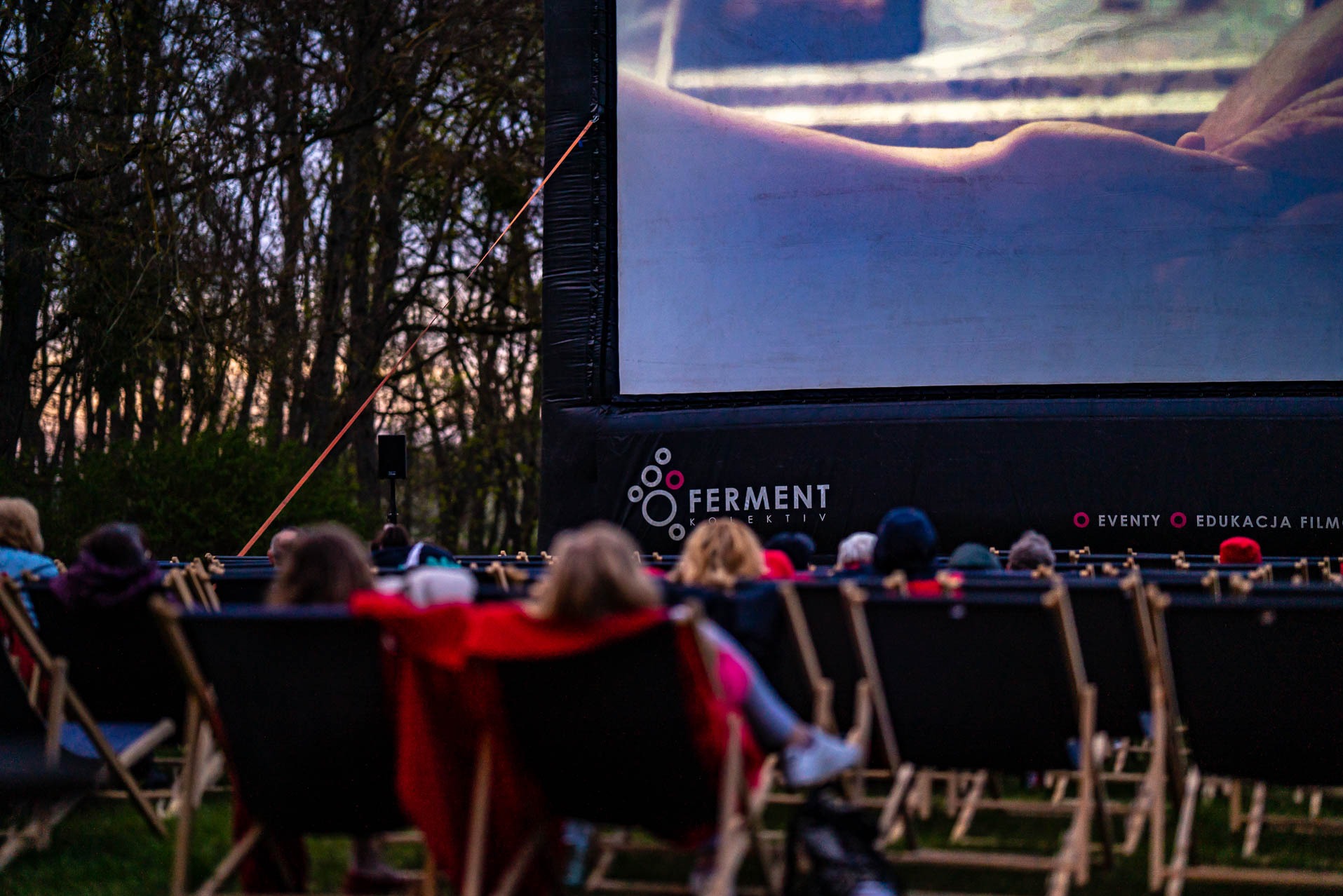 Kino plenerowe z widownią na leżakach, wyświetlany film na dmuchanym ekranie z logo firmy eventowej, drzewa w tle, wieczorny klimat.
