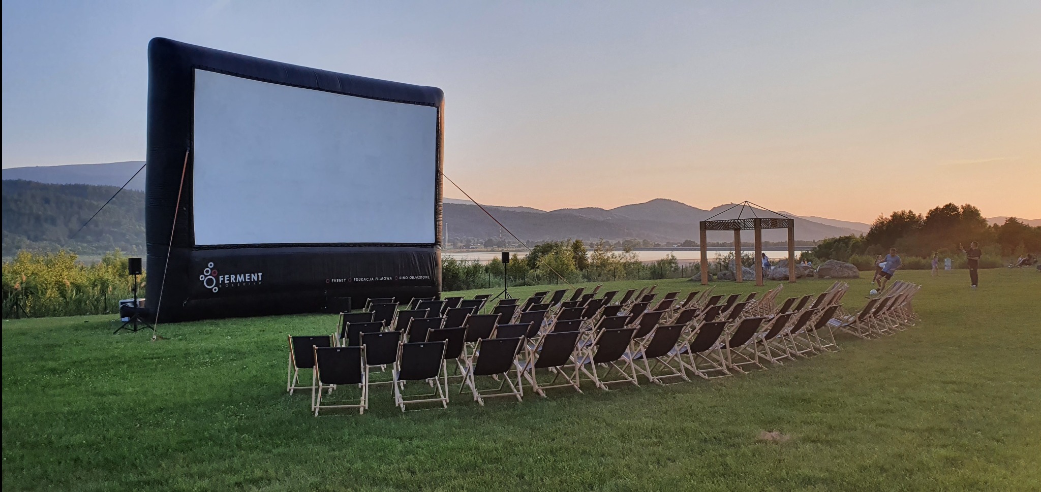 Kino plenerowe z dmuchanym ekranem i rzędami krzeseł na trawie, w tle jezioro i góry o zmierzchu. Widoczny logotyp 'Ferment O!lskikv'.