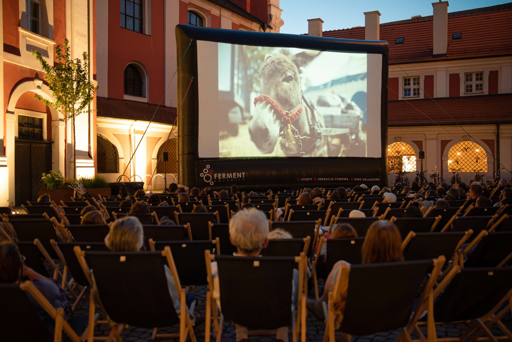 Kino plenerowe w zabytkowym otoczeniu: duży ekran z wyświetlanym filmem, rzędy leżaków z widzami, oświetlony budynek w tle. Wieczorny seans filmowy.