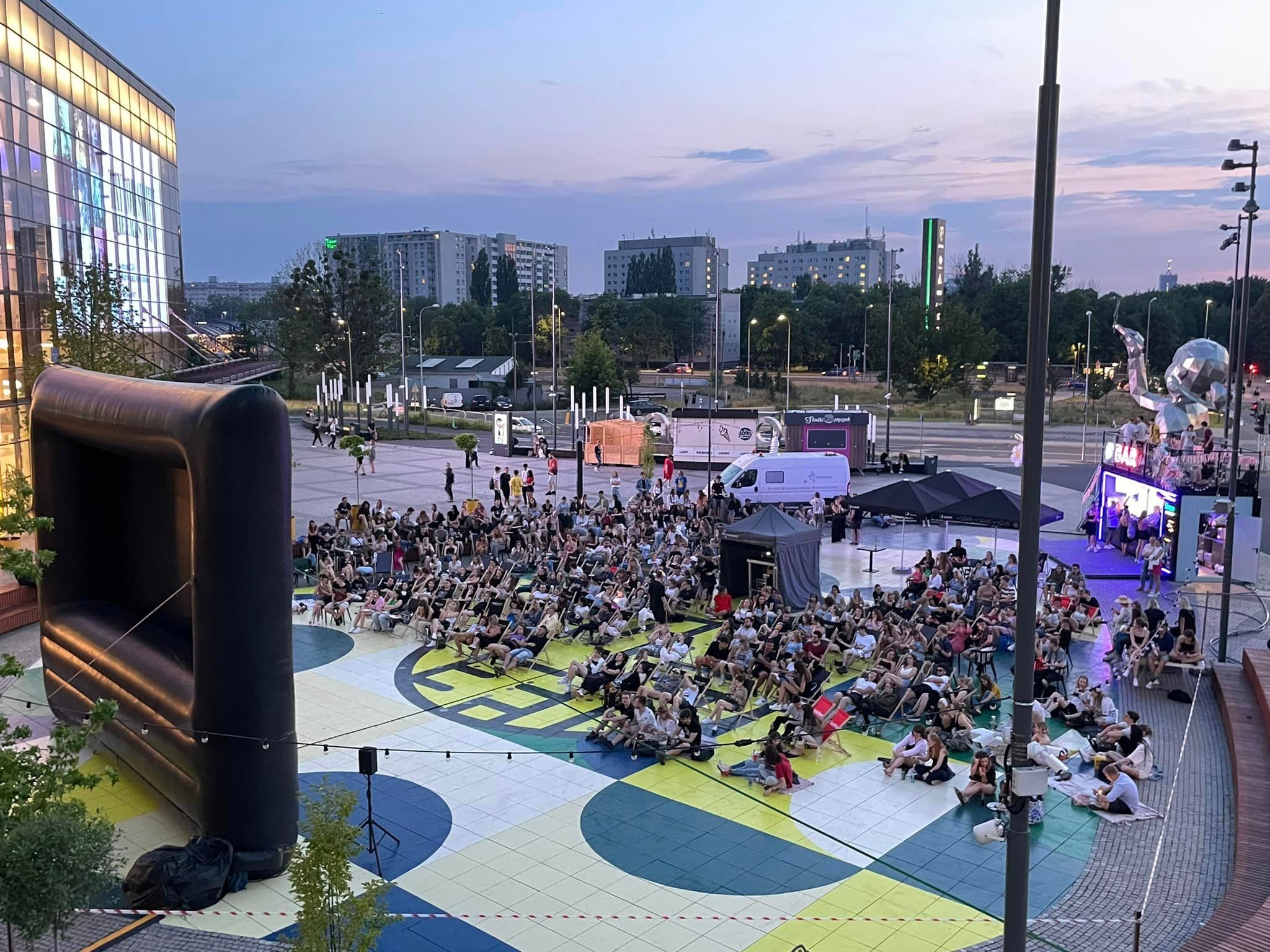 Plenerowe kino letnie z dużym dmuchanym ekranem, widownią na leżakach i sceną w tle, zorganizowane na placu miejskim o zmierzchu.