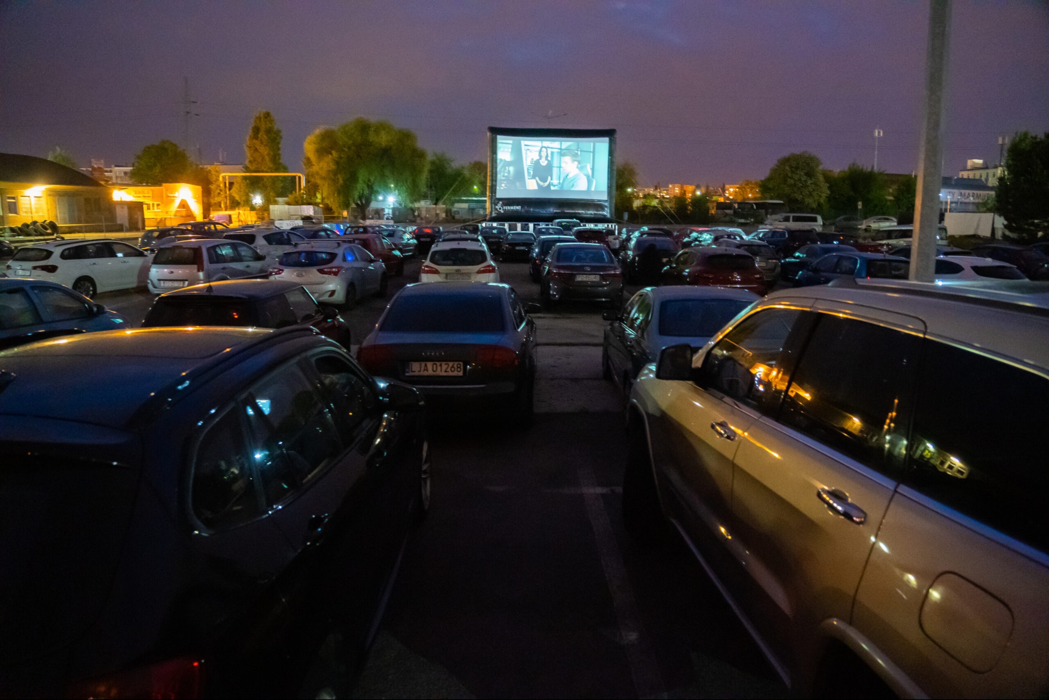 Nocne kino samochodowe: rzędy aut skierowane do ekranu z wyświetlanym filmem, oświetlonym przez reflektory. Atmosfera wieczornego wydarzenia.