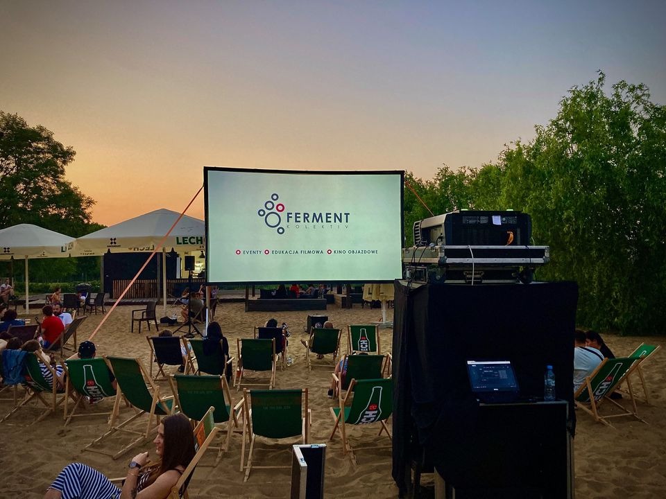 Kino plenerowe na plaży z ekranem 'Ferment Kolektyw', rzędami leżaków i projektorem. Widzowie siedzą, w tle parasole i drzewa, zachodzące słońce.