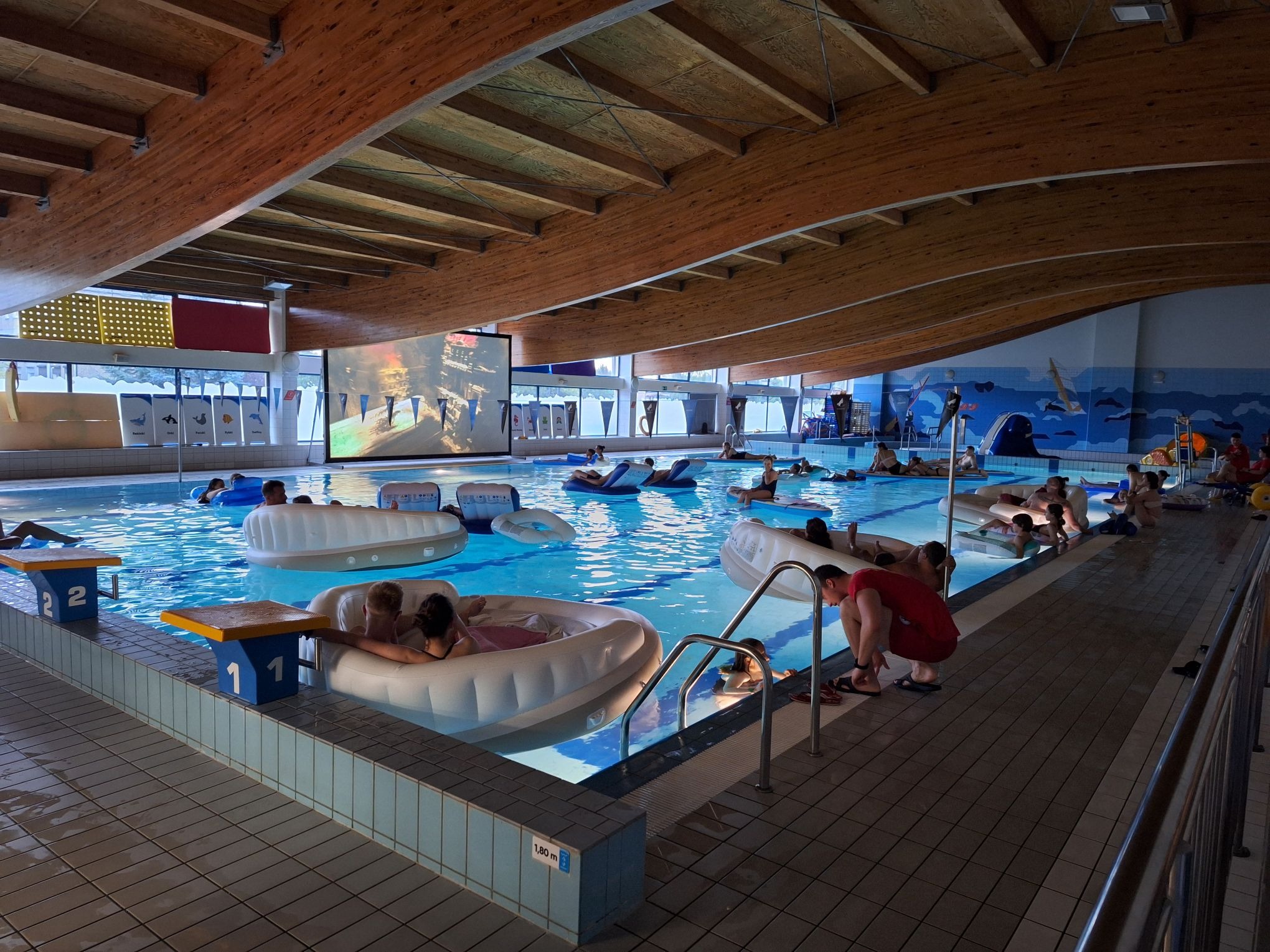 Basen z osobami na dmuchanych materacach podczas projekcji filmu. Drewniany sufit, niebieskie płytki i woda. Atrakcja eventowa w aquaparku.