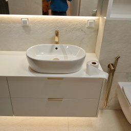 Remonty-wykonczenia wnetrz - Nowoczesna łazienka z białą umywalką nablatową, złotą armaturą i podświetlaną szafką. W lustrze odbija się osoba robiąca zdjęcie.