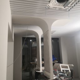 Remonty-wykonczenia wnetrz - Wnętrze w trakcie prac wykończeniowych: widoczne kolumny z surowego betonu, podwieszany sufit lamelowy i klimatyzacja. W tle wózek z narzędziami.