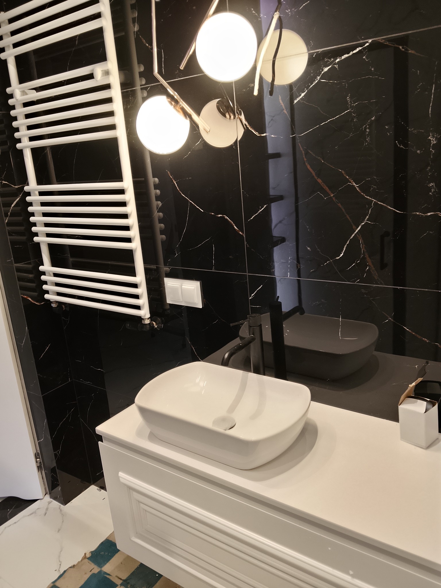 Nowoczesna łazienka z czarnymi płytkami imitującymi marmur, białą umywalką nablatową i grzejnikiem drabinkowym. Widoczne oświetlenie sufitowe i fragment podłogi.