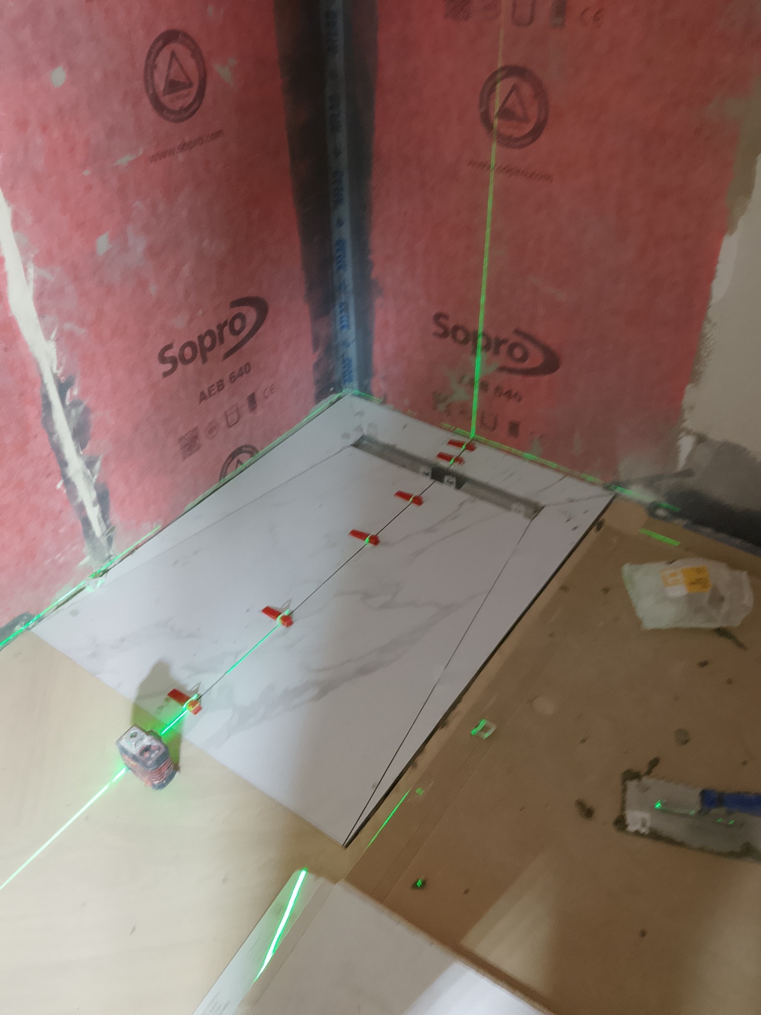 Układanie białych płytek w łazience z użyciem poziomicy laserowej. Widoczne czerwone kliny dystansowe i hydroizolacja Sopro na ścianach. Prace glazurnicze w toku.