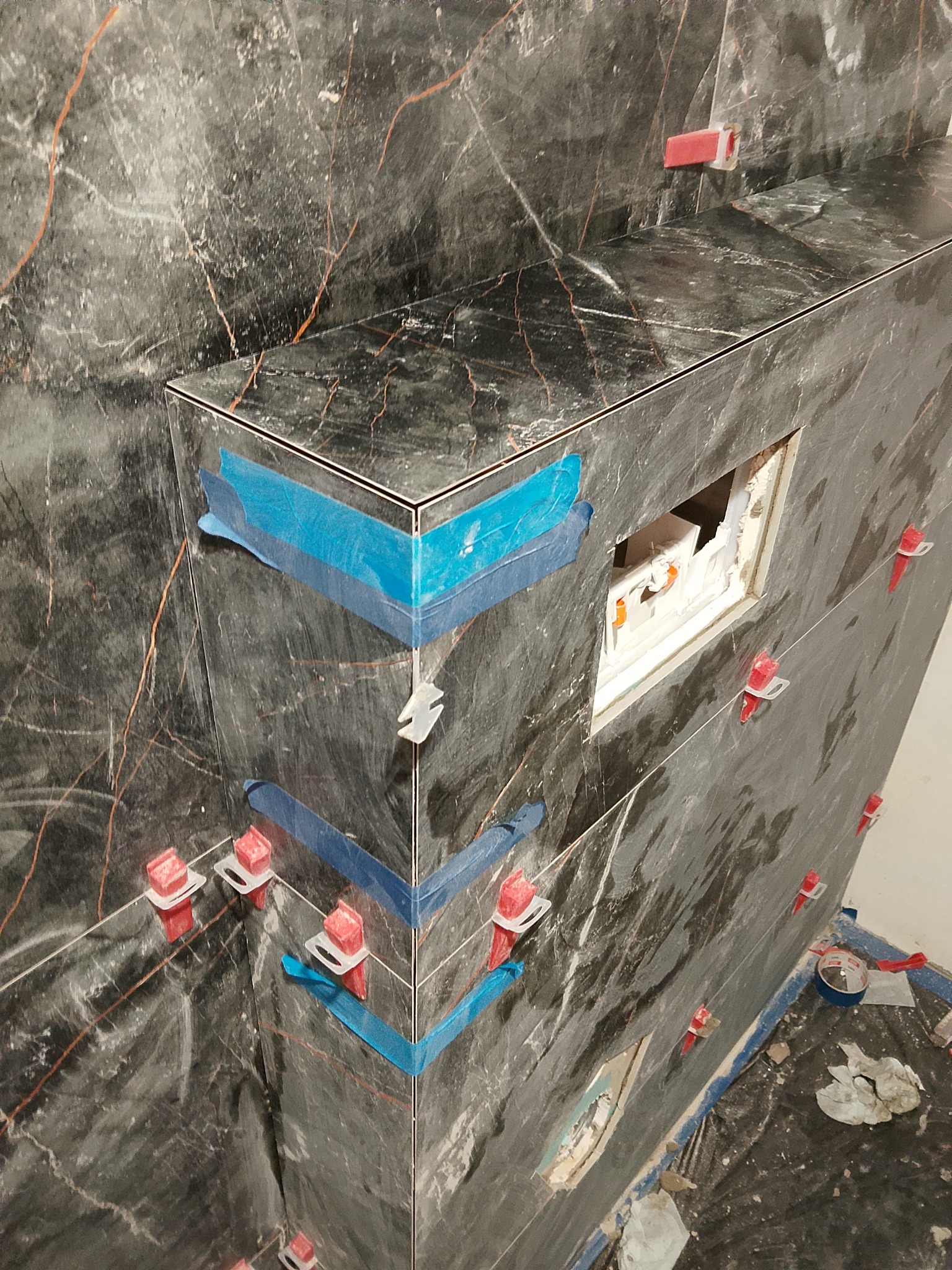 Montaż płytek imitujących marmur w łazience, z użyciem poziomujących klipsów i taśmy malarskiej. Widoczny otwór na instalację sanitarną.