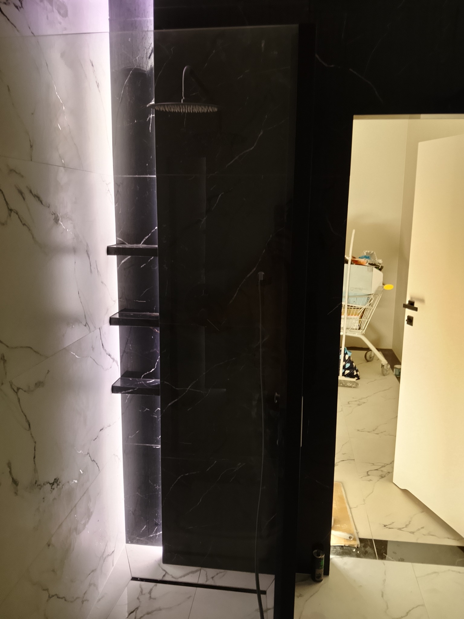 Nowoczesna kabina prysznicowa z czarnymi i białymi płytkami imitującymi marmur, podświetlane półki, widok na wnętrze łazienki z wózkiem transportowym.