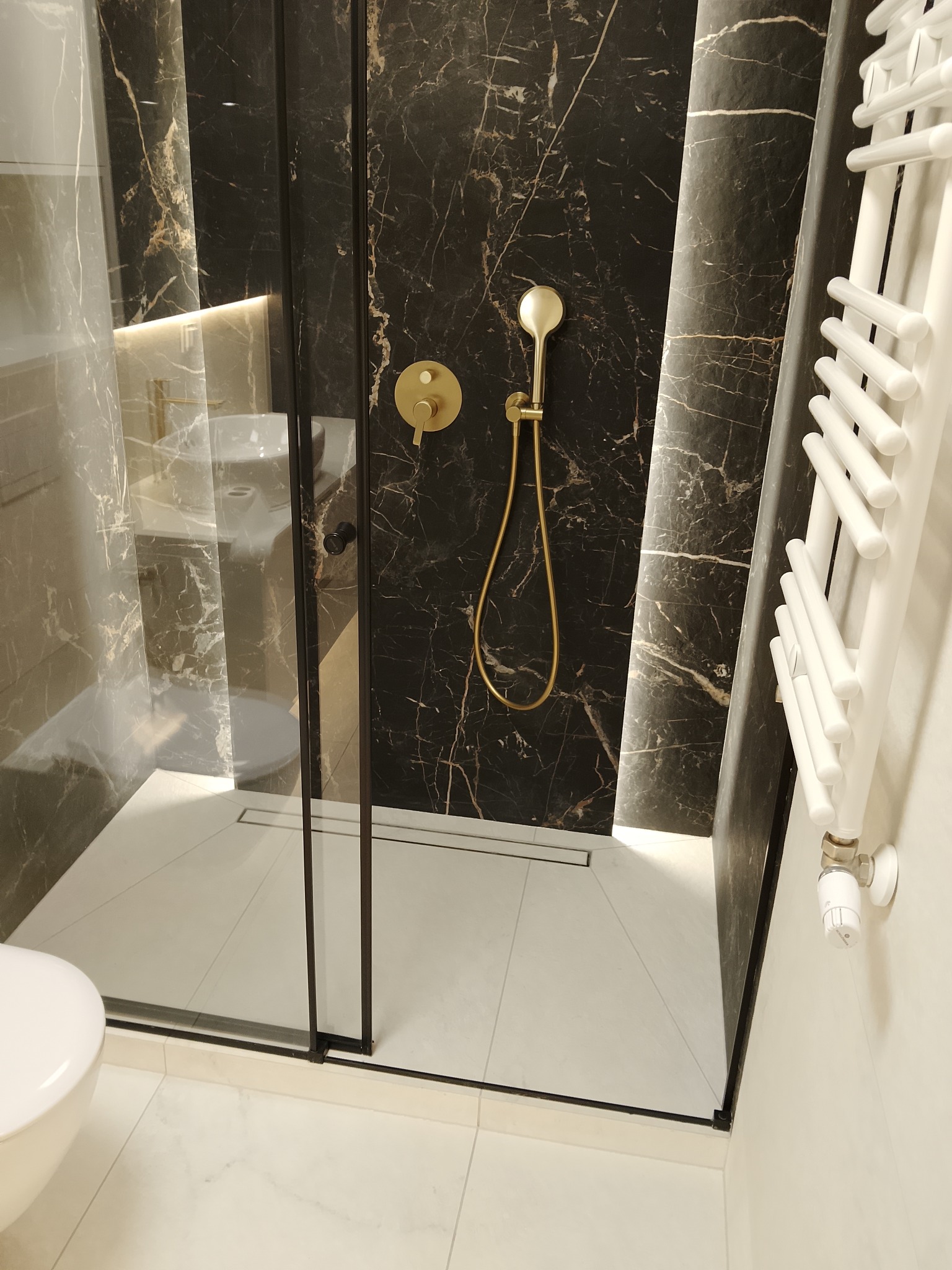 Nowoczesna kabina prysznicowa z czarnym marmurem, złotą armaturą i białym grzejnikiem drabinkowym. Minimalistyczny design, eleganckie wykończenie.