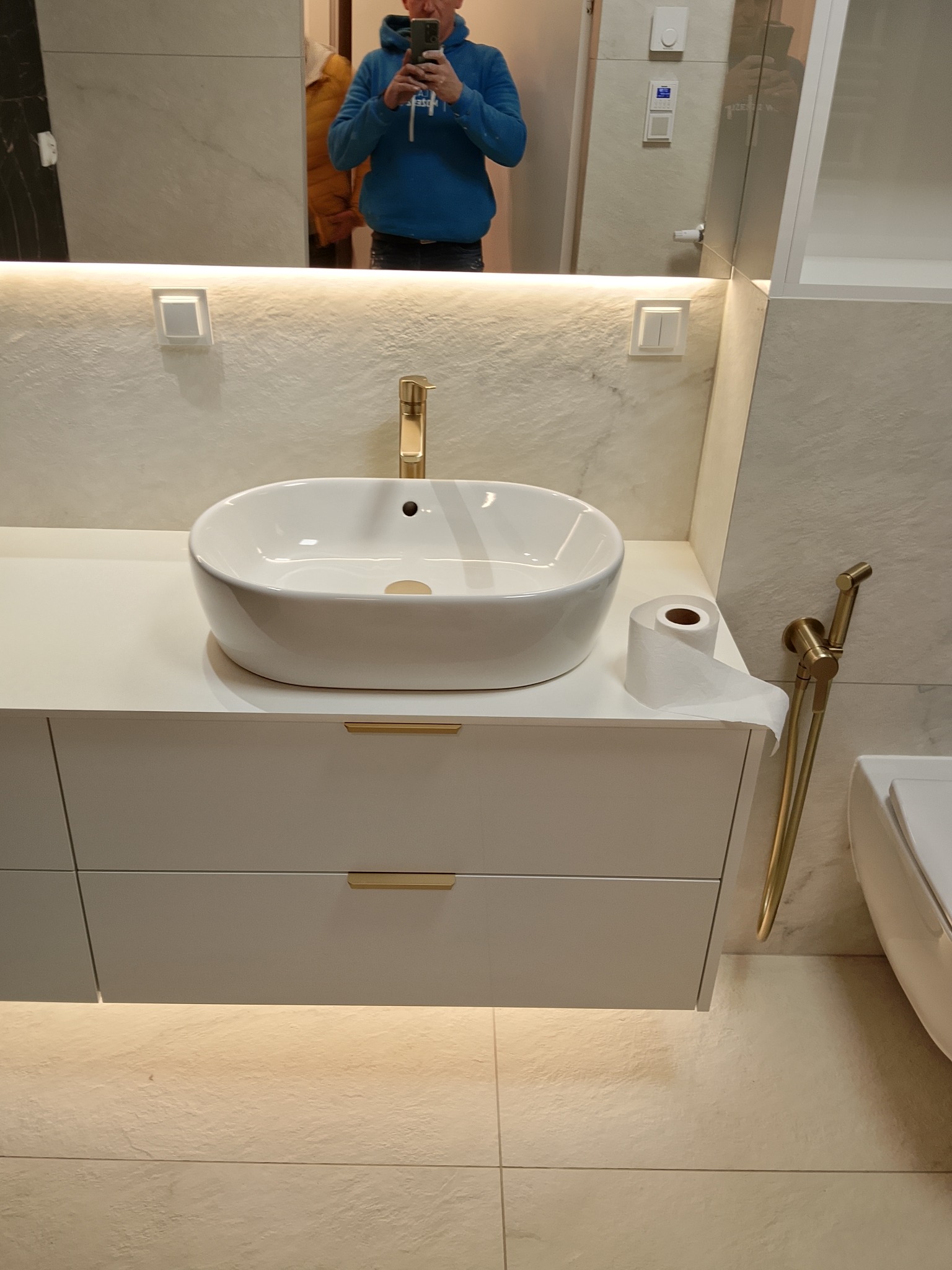 Nowoczesna łazienka z białą umywalką nablatową, złotą armaturą i podświetlaną szafką. W lustrze odbija się osoba robiąca zdjęcie.