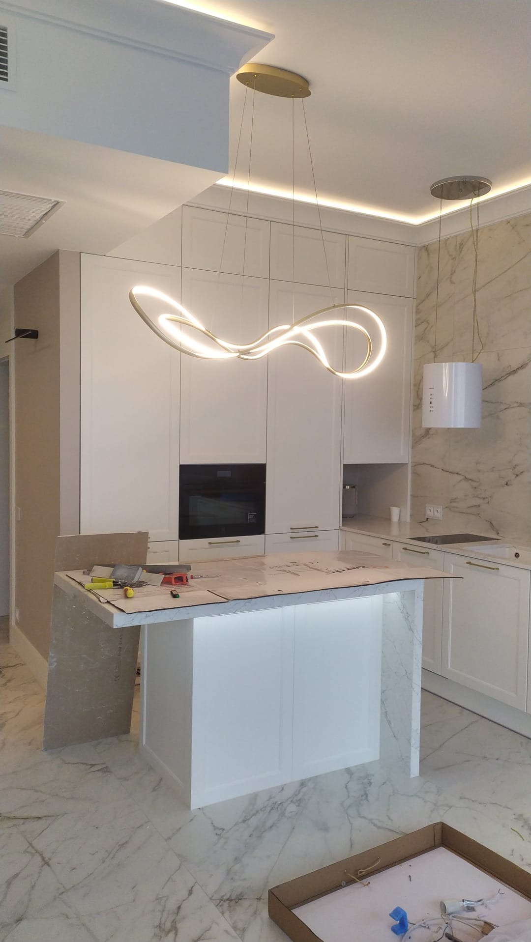 Biała kuchnia z wyspą i nowoczesnym oświetleniem LED. Na blacie wyspy narzędzia, co sugeruje trwający montaż. Marmurowe wykończenie ścian i podłogi dodaje elegancji.