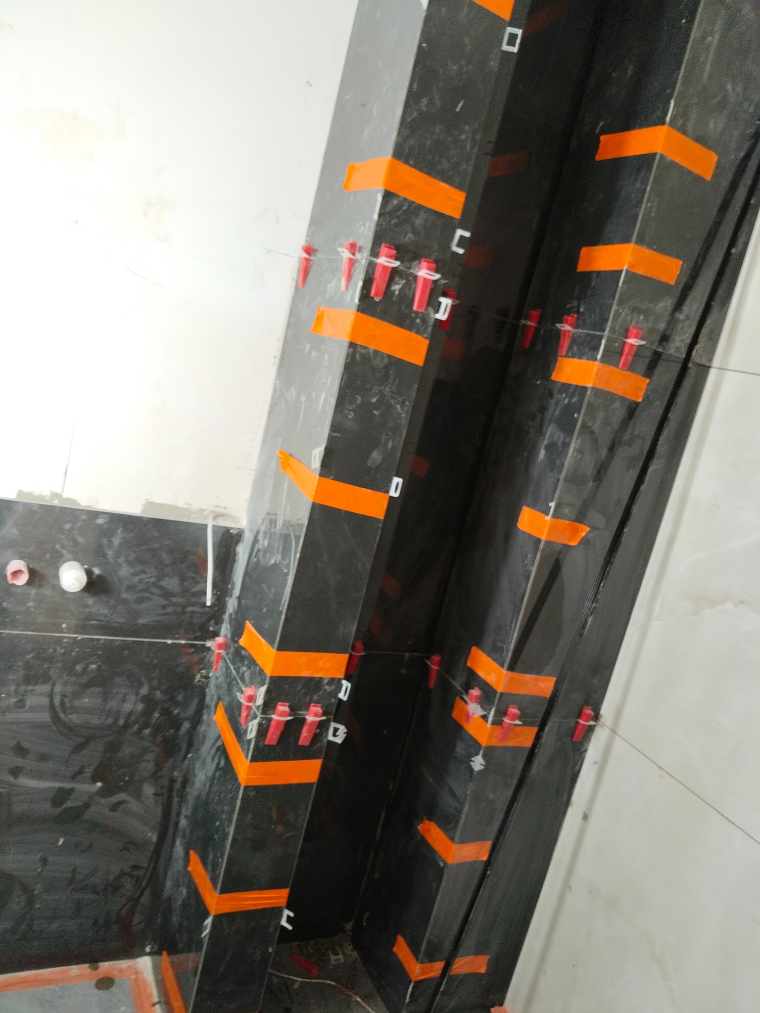 Układanie czarnych płytek na filarze z użyciem poziomicy i klipsów w Wołczkowie. Prace glazurnicze w trakcie realizacji, widoczne pomarańczowe taśmy i elementy mocujące.