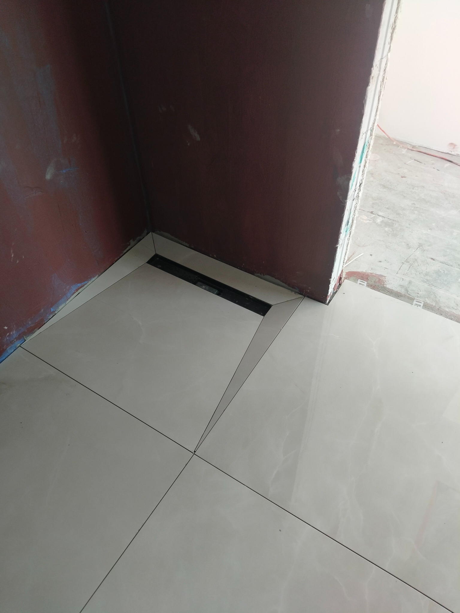 Ułożone, jasne płytki podłogowe z odpływem liniowym w rogu pomieszczenia. Widoczne niedomalowane ściany w odcieniach brązu i fragment surowego stanu wykończenia.