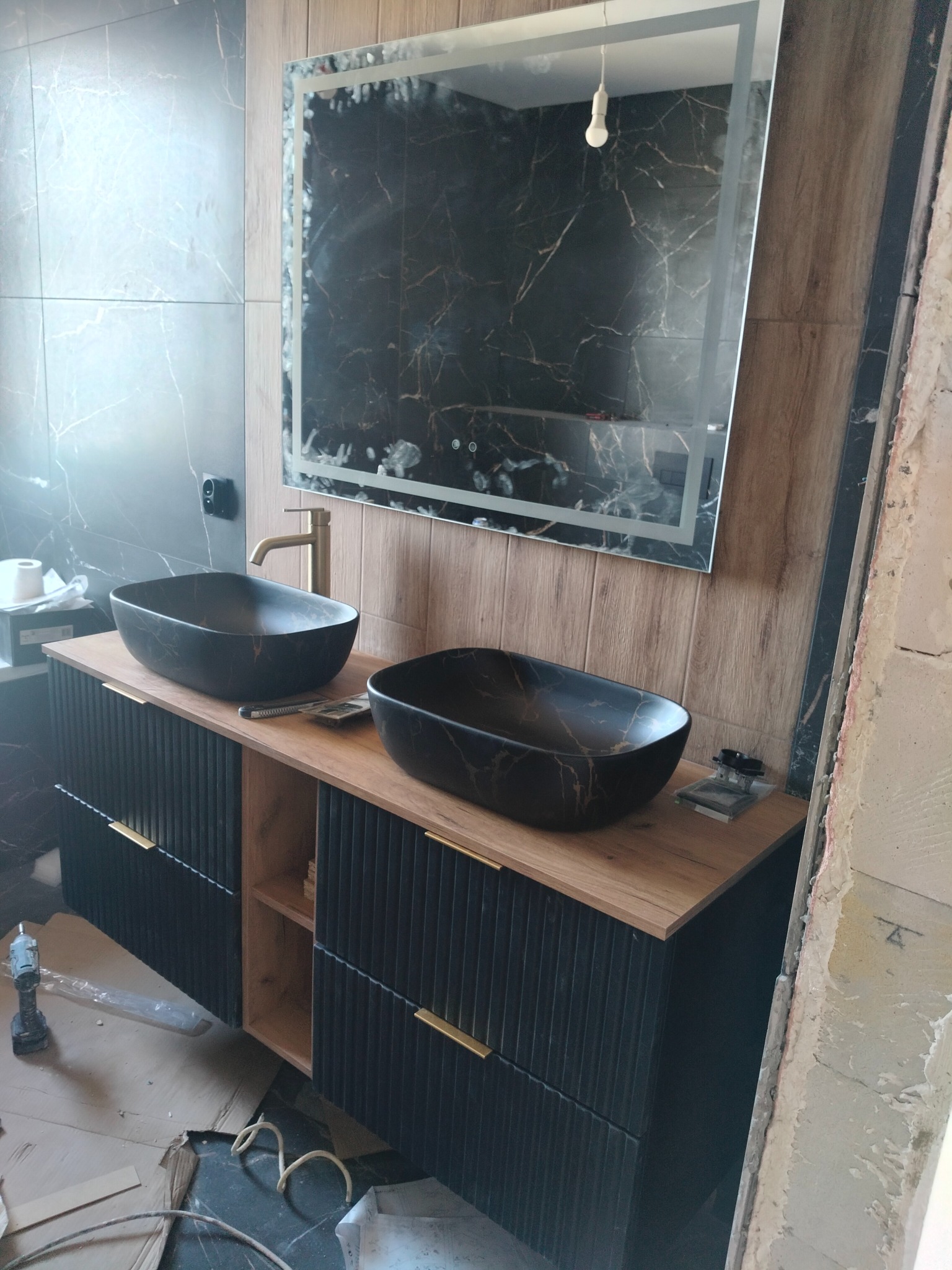 Nowoczesna łazienka z dwoma umywalkami w kolorze czarnym, złotą armaturą i podświetlanym lustrem. Widoczne elementy wykończenia i narzędzia.