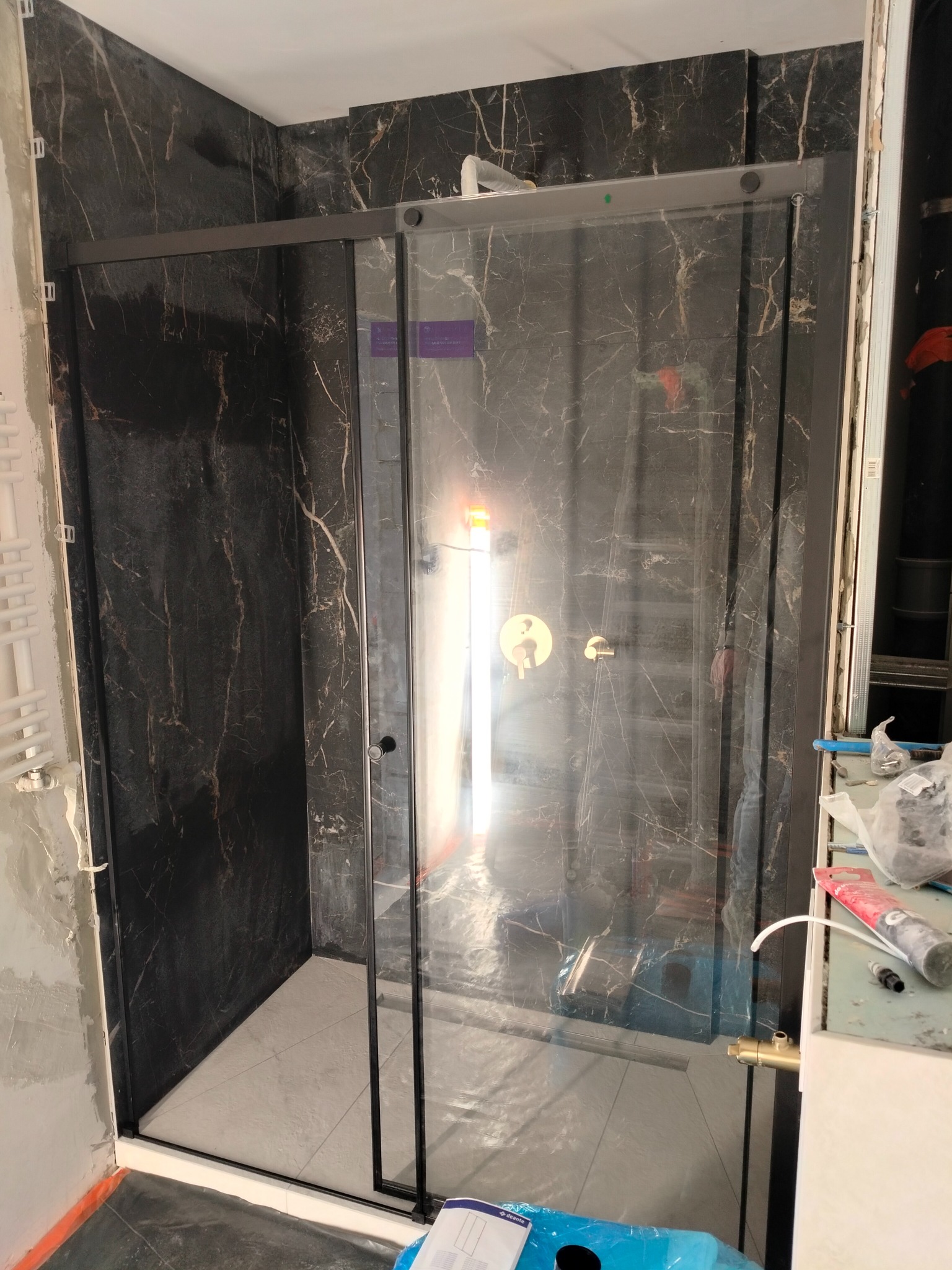 Kabina prysznicowa z czarnymi profilami i marmurowymi płytkami w trakcie montażu. Widoczne narzędzia i materiały budowlane. Ujęcie z bliska, podkreślające detale wykończenia.