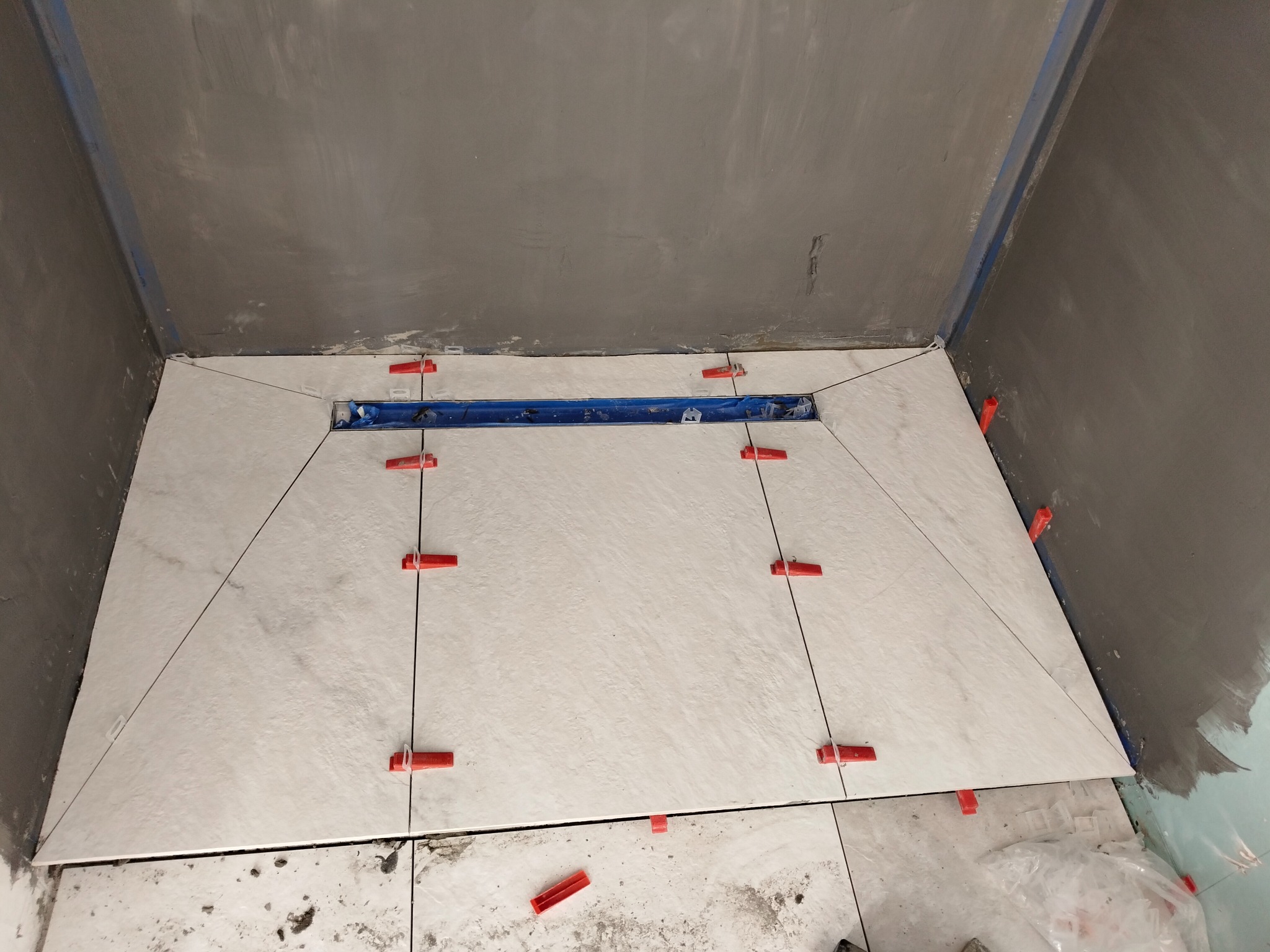 Układanie jasnych płytek podłogowych w łazience z odpływem liniowym, widoczne czerwone klipsy poziomujące. Ściany surowe, przygotowane do dalszych prac.