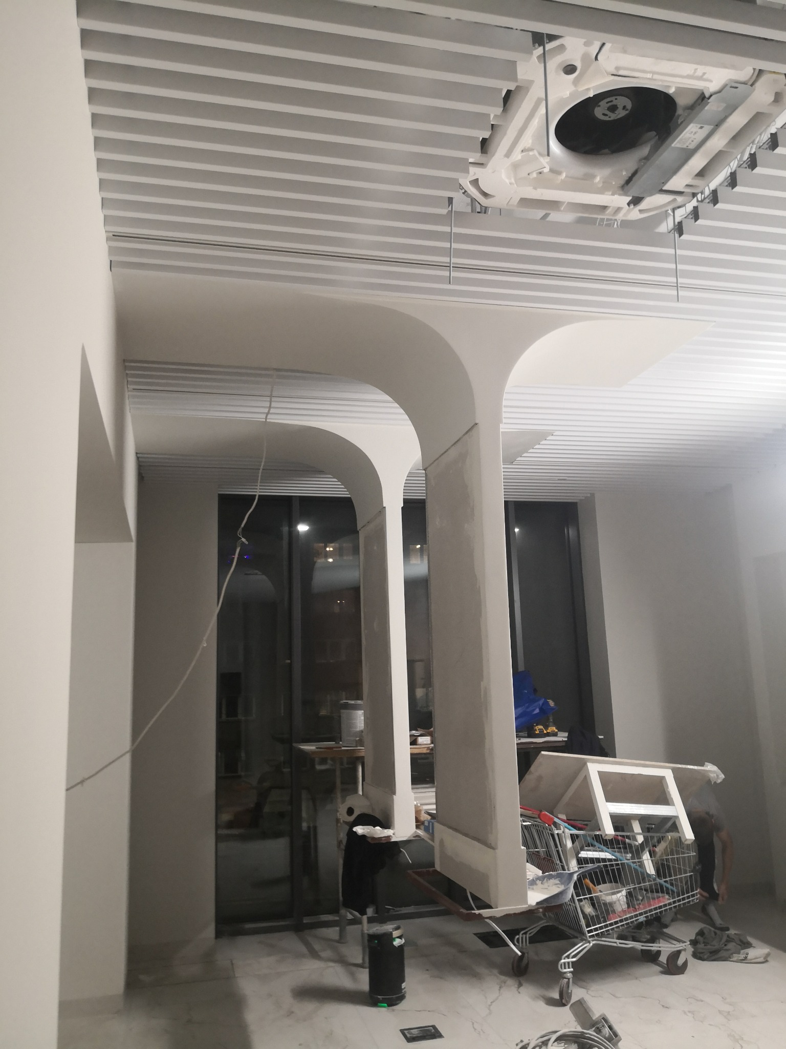 Wnętrze w trakcie prac wykończeniowych: widoczne kolumny z surowego betonu, podwieszany sufit lamelowy i klimatyzacja. W tle wózek z narzędziami.