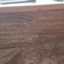 Laki - Fragment ściany z drewnianych bali z widocznym ociepleniem ze słomy i gliny, częściowo usuniętym, ukazującym konstrukcję. Narzędzia na górze kadru.