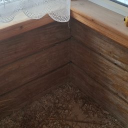 Laki - Fragment drewnianego okna z firanką i widokiem na zewnątrz, ukazujący stan podłogi przed pracami wykończeniowymi. Widoczne resztki gruzu i słomy.